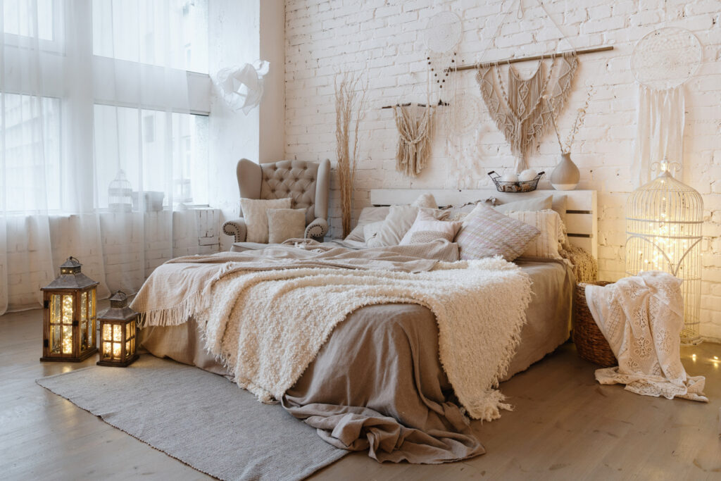 Boční pohled na krásnou ložnici s plédem a polštáři na pohodlné posteli, domácí výzdobou a polštáři na měkkém křesle v bílém interiérovém designu v bohémském stylu