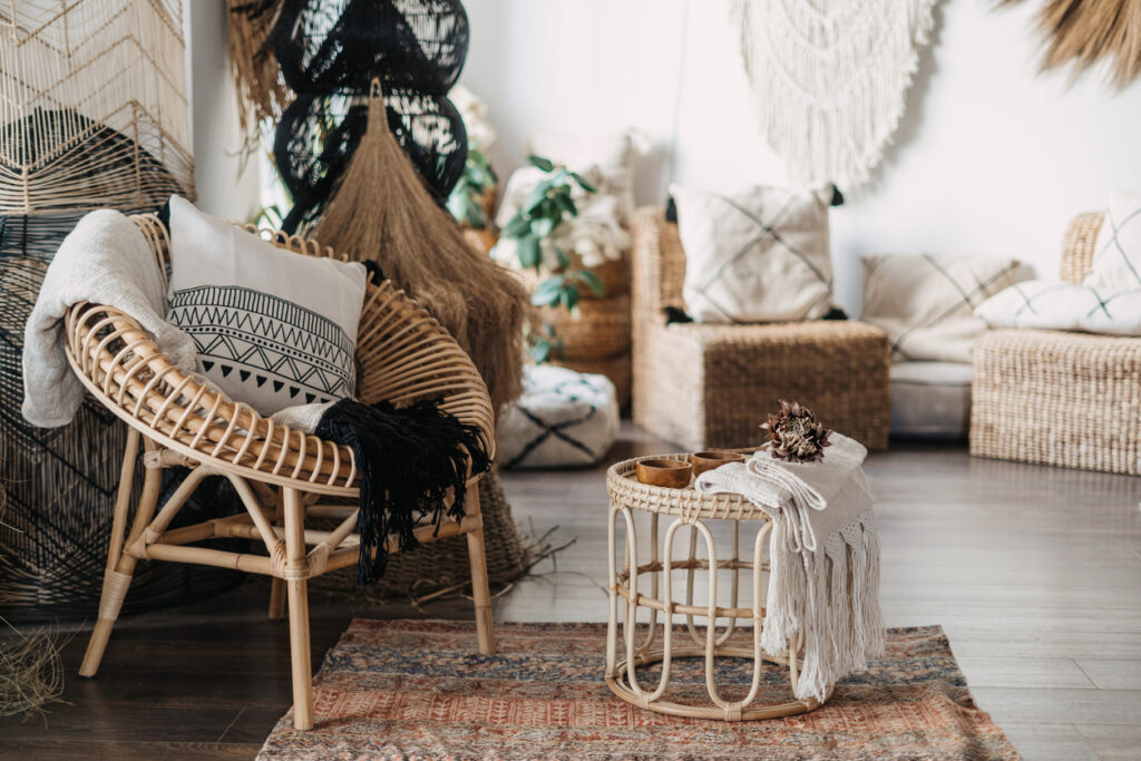 Pohodlný obývací pokoj s etnickým interiérovým designem v bohémském stylu. Byt s domácí výzdobou, ratanový nábytek, křeslo s polštáři, bambusový konferenční stolek, koberec na dřevěné podlaze